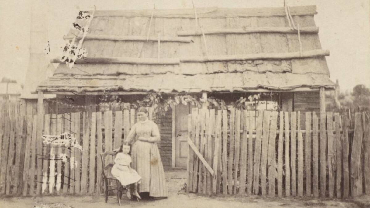 Woman and seated child outside a weatherboard cottage with bark roof and paling fence in Hill End, circuit 1872. Picture from the Holtermann Collection of Photographs via the State Library of NSW.